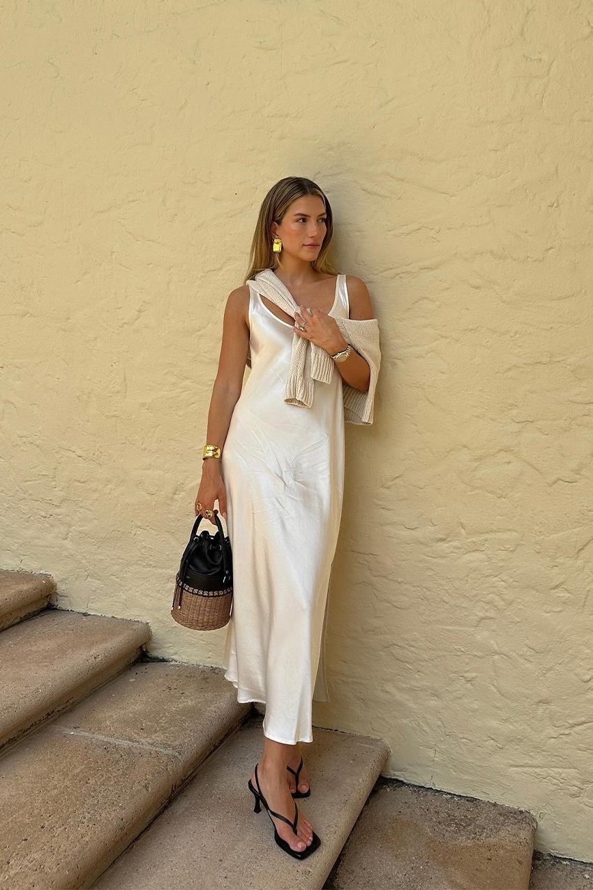 White outfit idea for summer is shown in a photo of a woman standing outside on stairs wearing gold earrings, a white slip dress, a white cardigan, a gold cuff bracelet, a raffia bucket bag, and black thong sandals