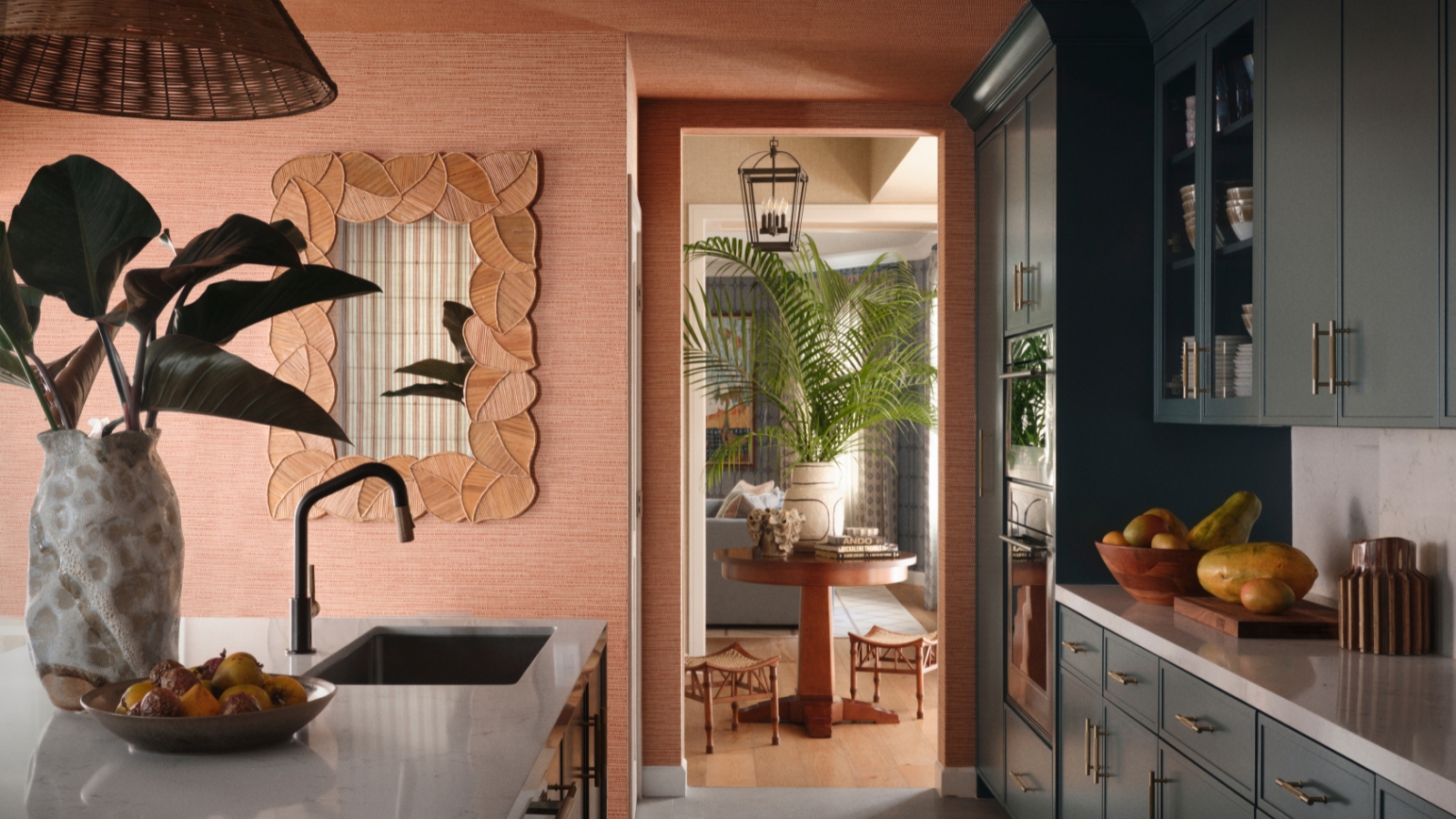 Kitchen with deep green cabinetry, terracotta raffia walls, white stone island, woven pendant and leaf-framed mirror.
