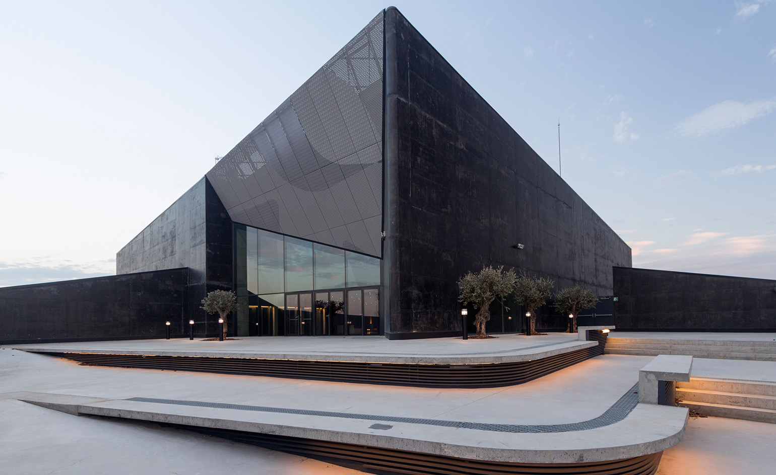 Exterior view of Caixaforum Cultural Exhibition Centre. A black building with dark reflective glasses. Grey steps leading to the building with mini trees at the entrance