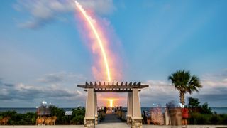 A bright streak of light curves through the blue sky behind a hut and pier.