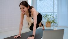 Woman exercises at home. She is on a black exercise mat, an open laptop is next to her, and she is wearing black athletic leggins and a white tank top