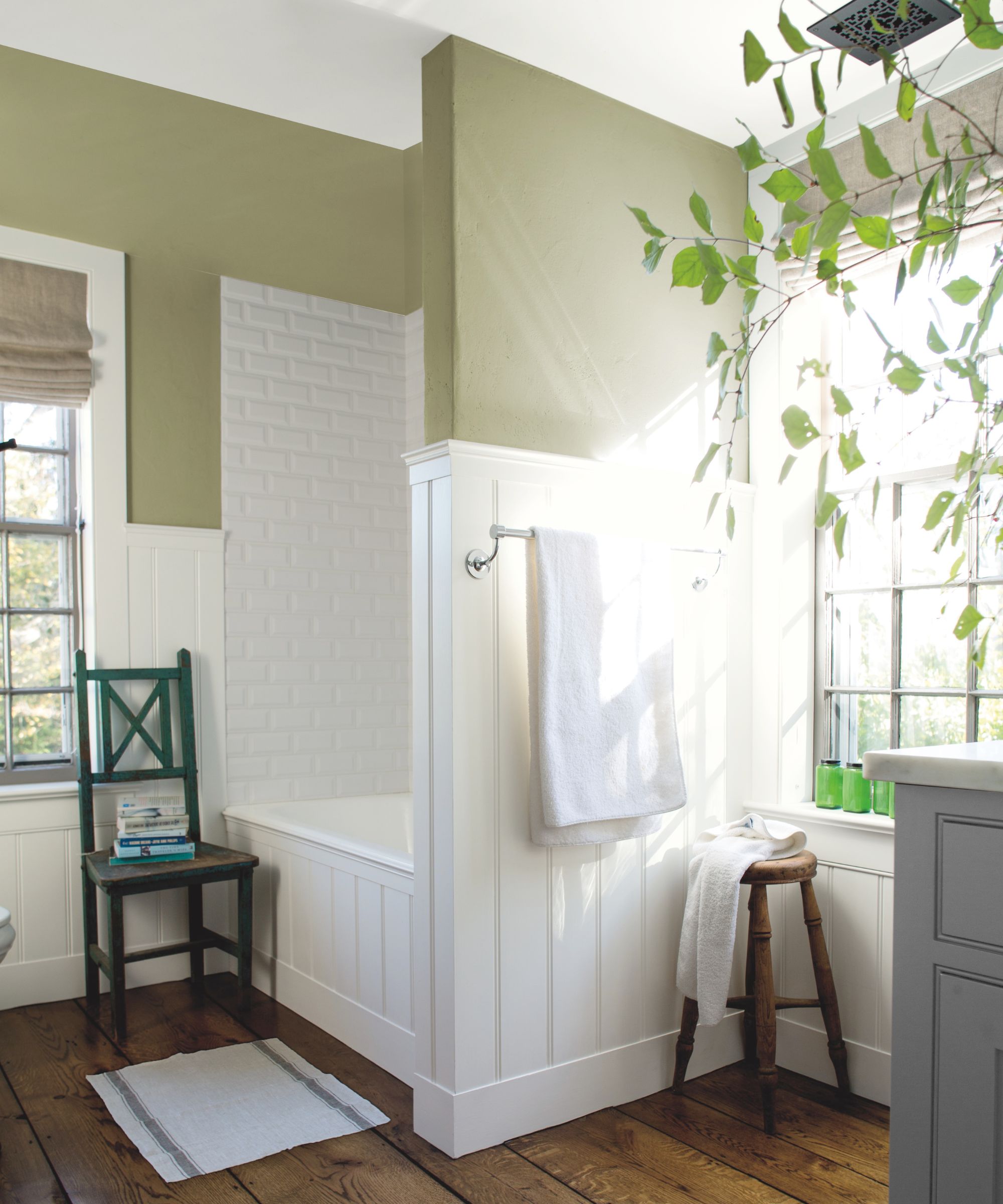 Bathroom with exposed wood floorboards, white wood panelled bath nook, wooden stool, green walls and plant by the sink