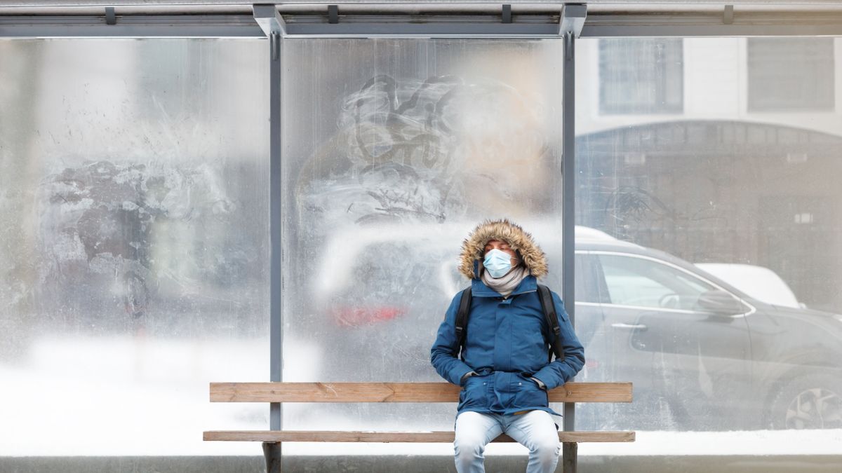 A man in a coat and mask sitting on a bench waiting for public transport.