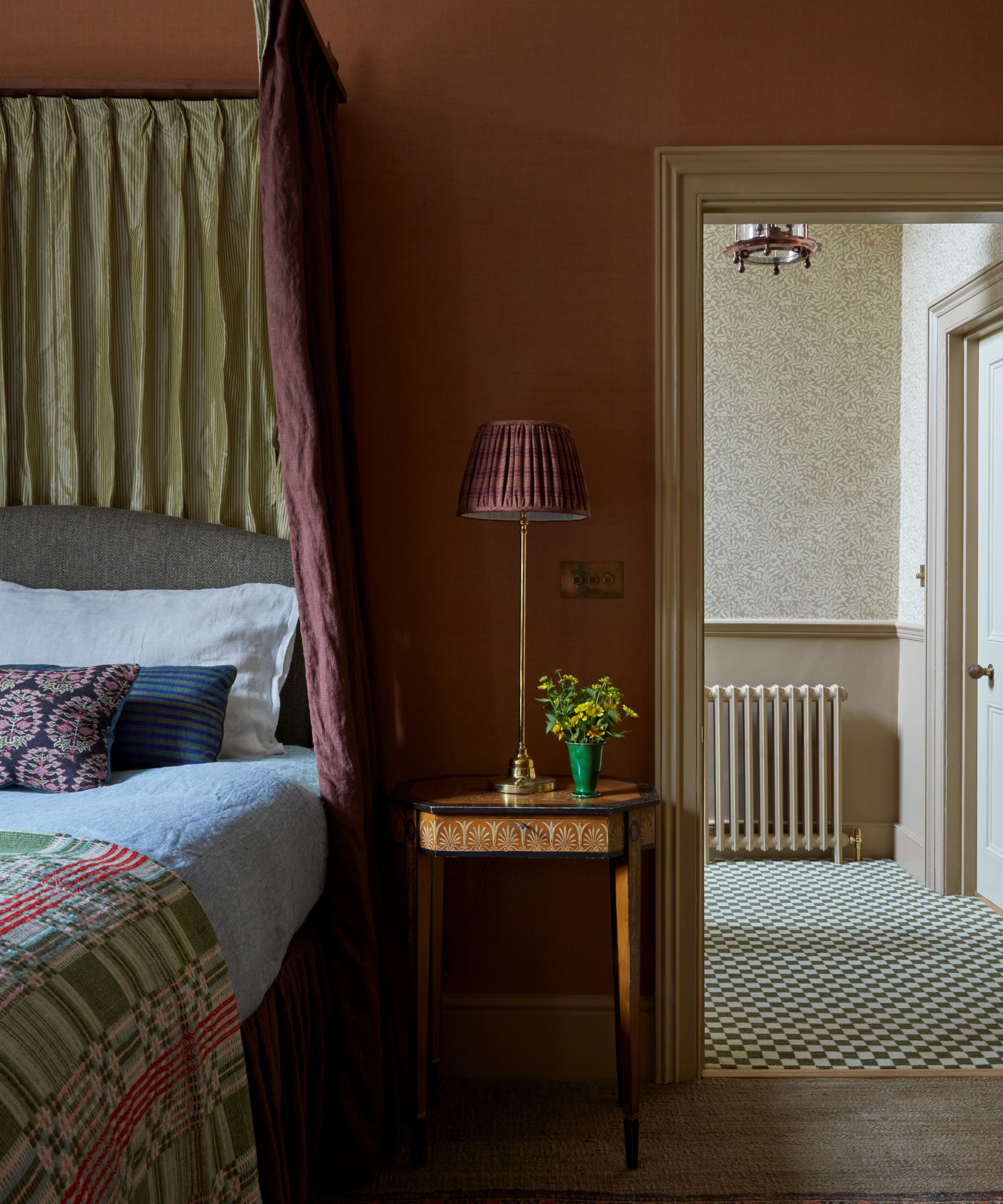 Bedroom with clay walls, wood side table, maroon silk canopy, white bedding with antique cushions looking out onto hallway