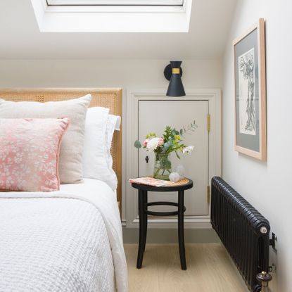 White bedroom with rattan headboard, black radiator and a black stool as a bedside table.