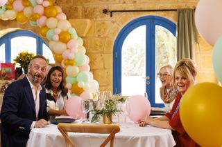 Dom (Steve Edge), Delphine (Julie Graham), Nancy (Lisa Maxwell) and Jean (Sally Lindsay) sit around a table in a fancy restaurant, decorated with balloons and flowers, and with a balloon arch in the distance. They all have expressions of delight on their faces.