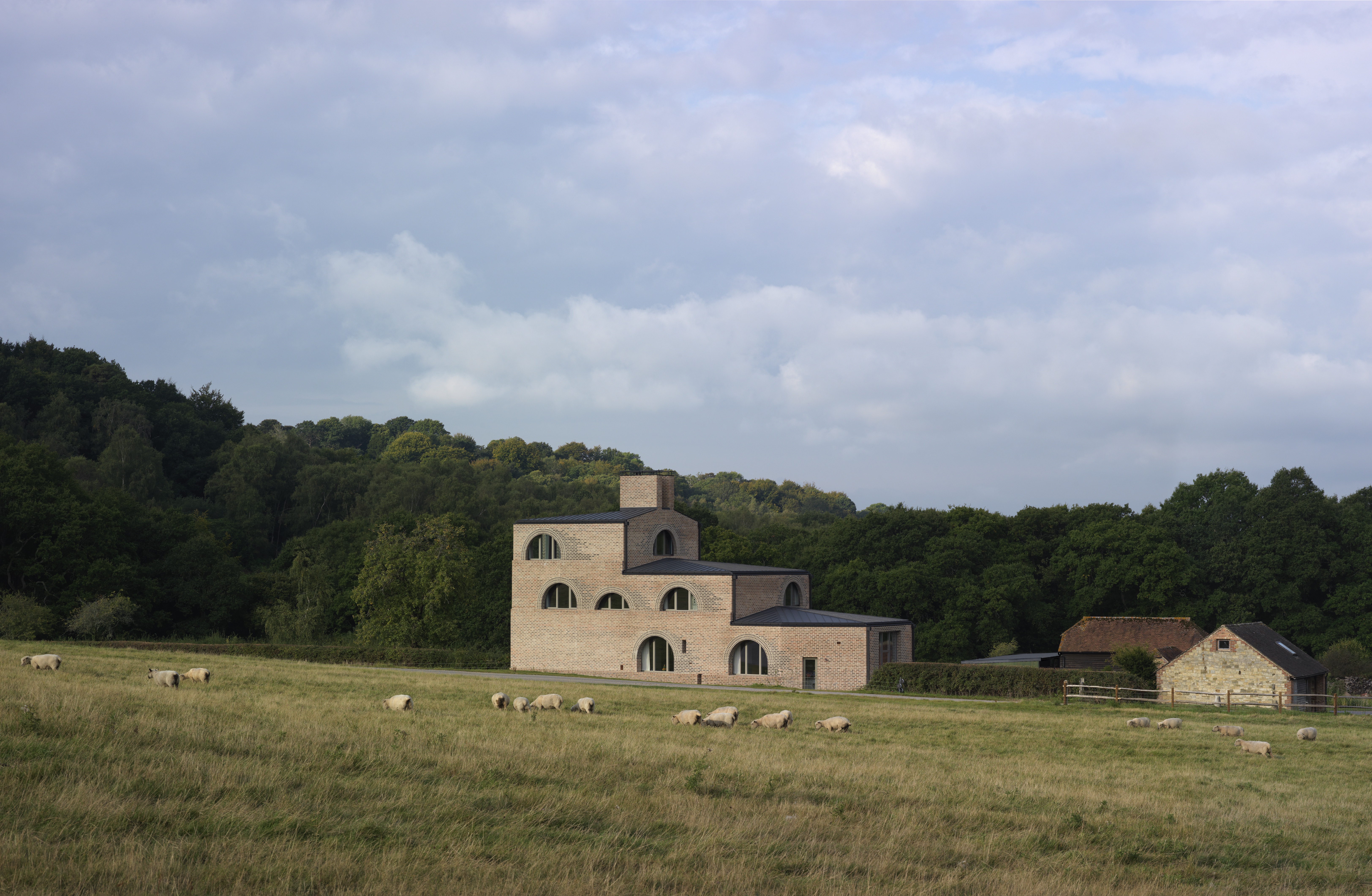 A large brick farm house with a stepped design with a field with sheep grazing in front of it and a forest behind it.