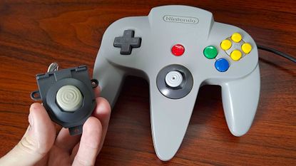Hand holding Hyperkin Hall Effect joystick next to grey Nintendo 64 controller on woodgrain desk.