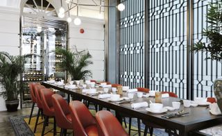 Interior view of the dining area at Empress featuring a wall with a geometric design, a long dark wood table with tableware, red chairs, black and grey pots with tall green plants, a grey and yellow patterned rug, a tall chrome coloured wine cooler and sphere lighting