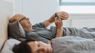 A man lying in bed looking at his wrist sleep tracker while his wife sleeps next to him