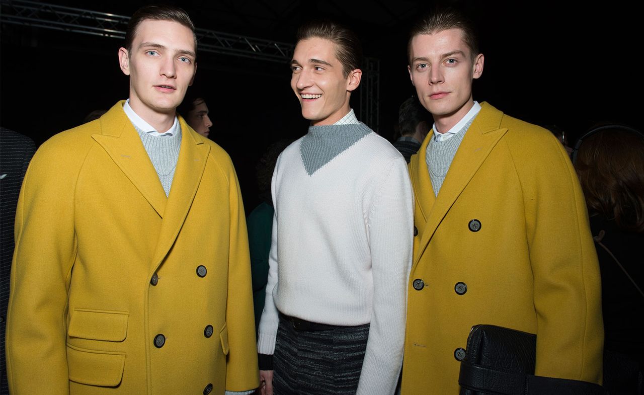 Three male models wearing looks from Canali&#039;s collection. They are wearing white shirts, white and grey jumpers, yellow jackets and striped grey trousers. One model is holding a bag