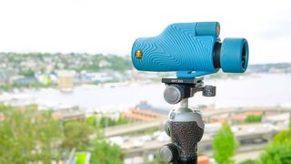 Nocs Field Tube Monocular in baby blue. Shown in a user's hand against a red background.