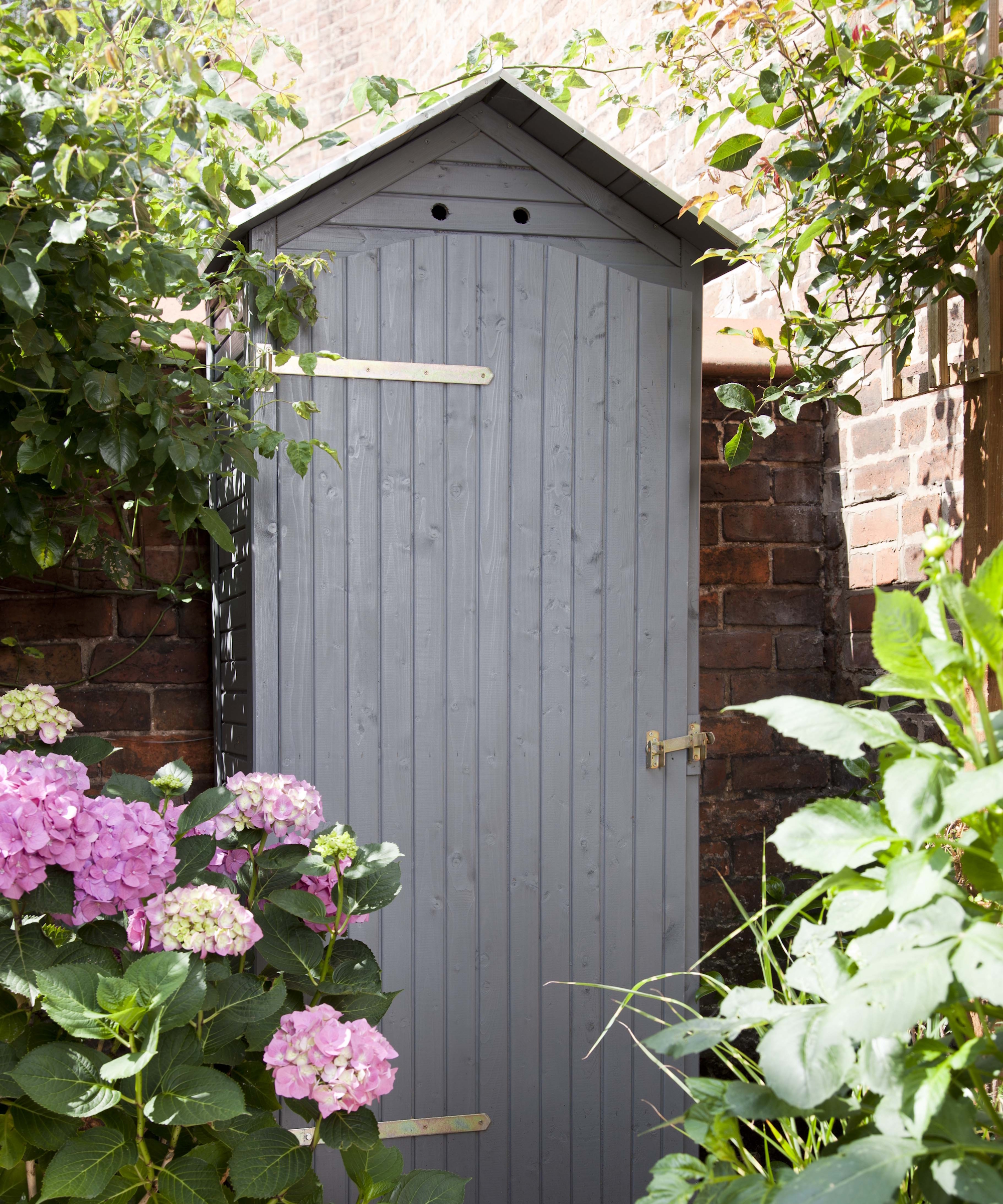 A small grey garden shed sits in the corner of a garden next to a brick wall