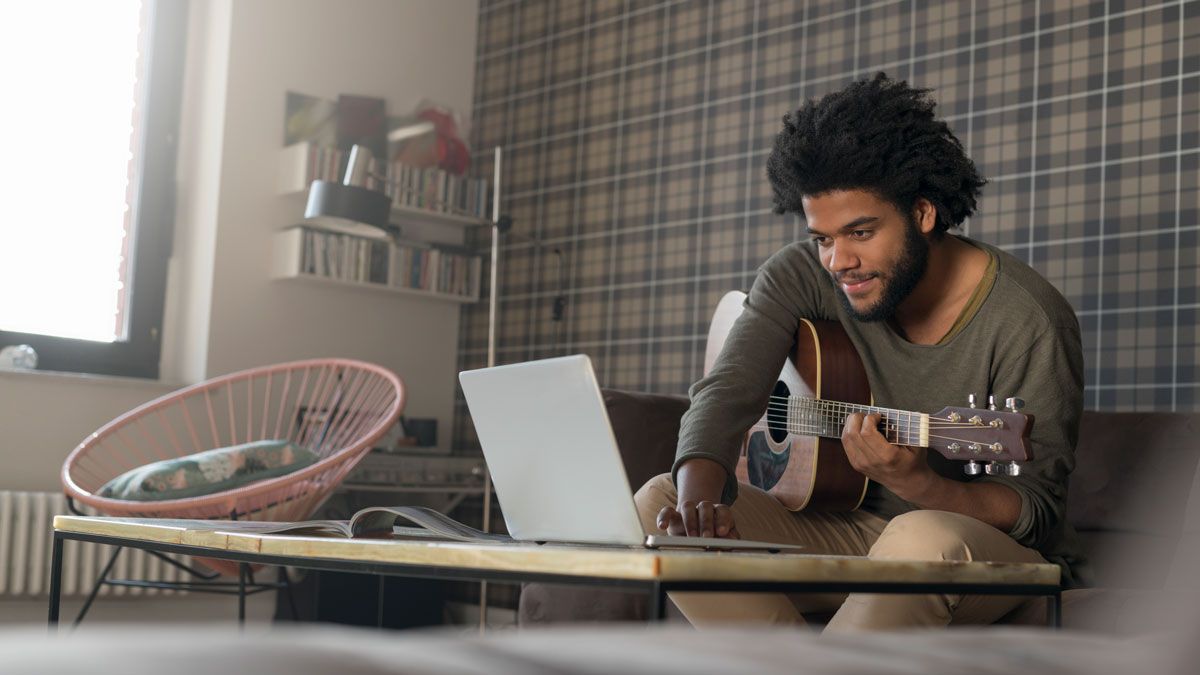 Man sitting in living room on sofa playing guitar in front of laptop