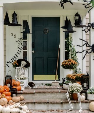Halloween door decor ideas with bat decorations on the walls, spiders climbing on the porch roof, seated skeletons, pumpkins and witches hats dangling from the roof