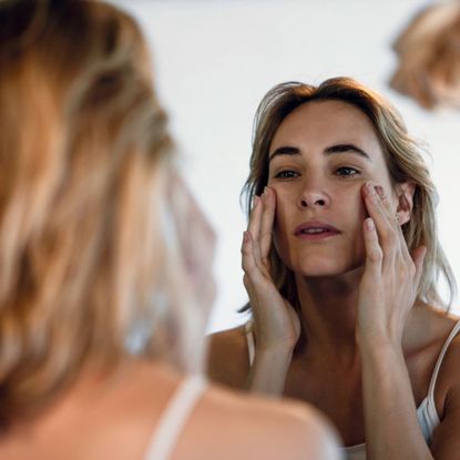 woman looking in the mirror with hands on her cheekbones - unexpected signs of ageing
