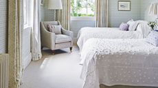 A bright twin bedroom with white bedding, green and white wallpaper, large windows with patterned curtains, and a large upholstered chair.