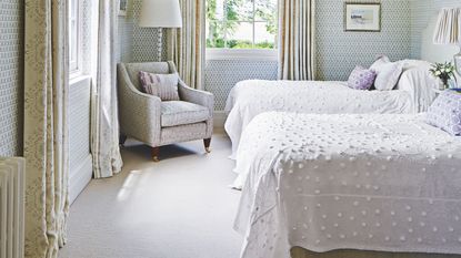 A bright twin bedroom with white bedding, green and white wallpaper, large windows with patterned curtains, and a large upholstered chair.