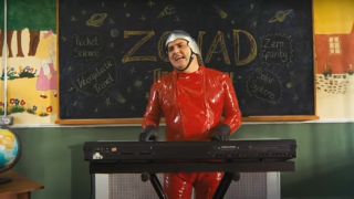 Simon Delaney in red leather singing at a keyboard while standing in front of a chalkboard in Zonad,