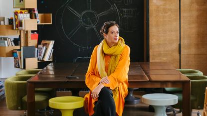 A woman wearing an orange wool dress, a yellow scarf and black trousers sit on a colorful stool in a room featuring modern decor.