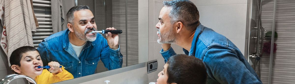 A picture of a father and young son brushing their teeth with electric toothbrushes while standing in front of a bathroom mirror