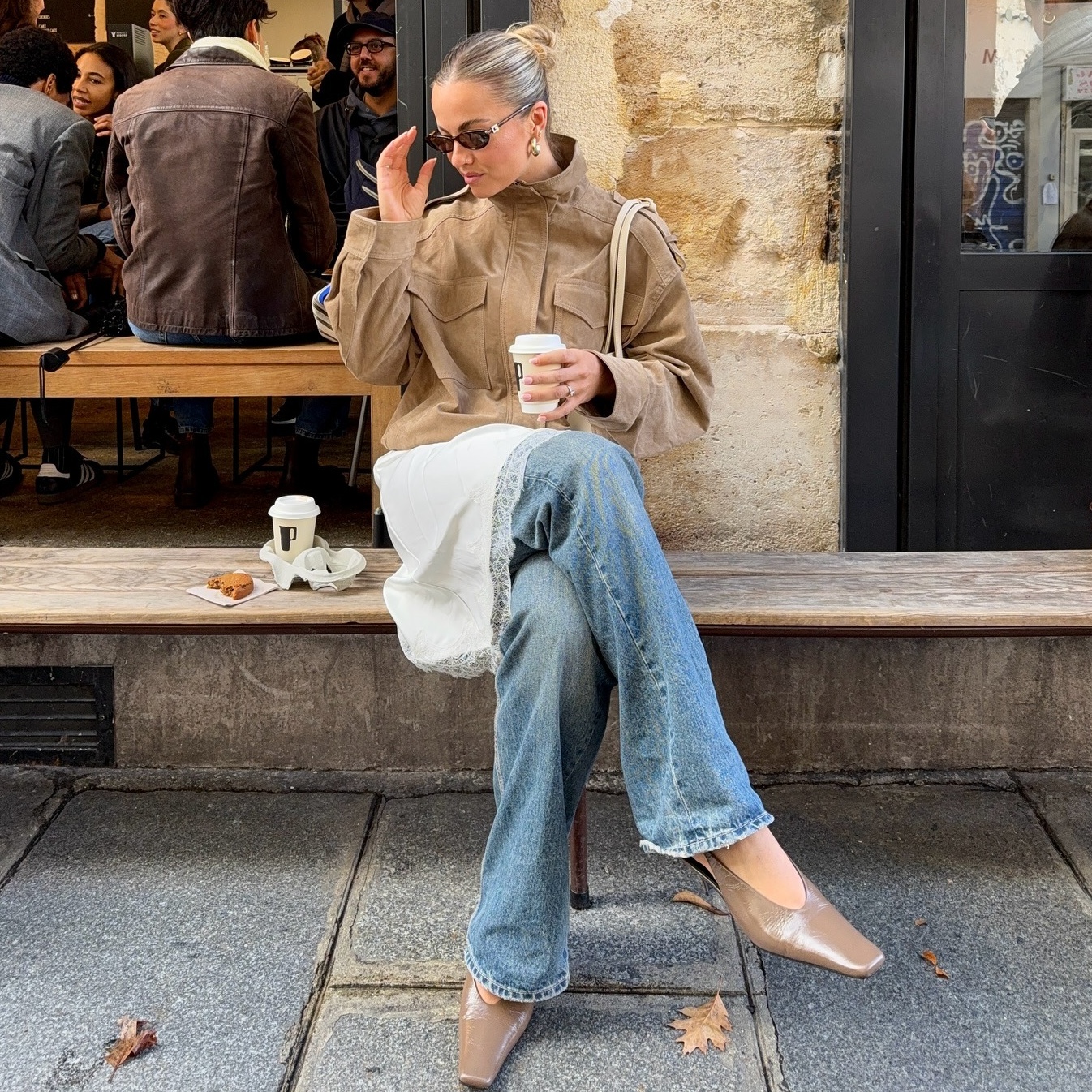 collage of style influencer Izzy Dilger sitting at a wood bench outside a cafe in Paris, France wearing sunglasses, a tan suede jacket with funnel neck, lace-trim dress, bootcut jeans, and neutral slingback kitten heels