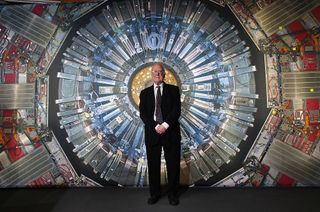 A man in a suit stands in front of an image of the Large Hadron Collider, which has lots of mechanical parts centered around a yellow middle device.