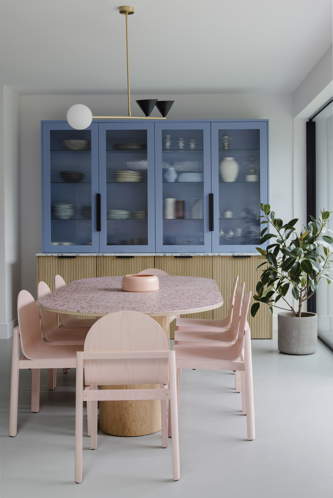 dining room with blue and timber cabinets displaying plates and crockery, pink stone and timber dining table, with pink dining chairs, a ficus in a pot, and gray polished floors