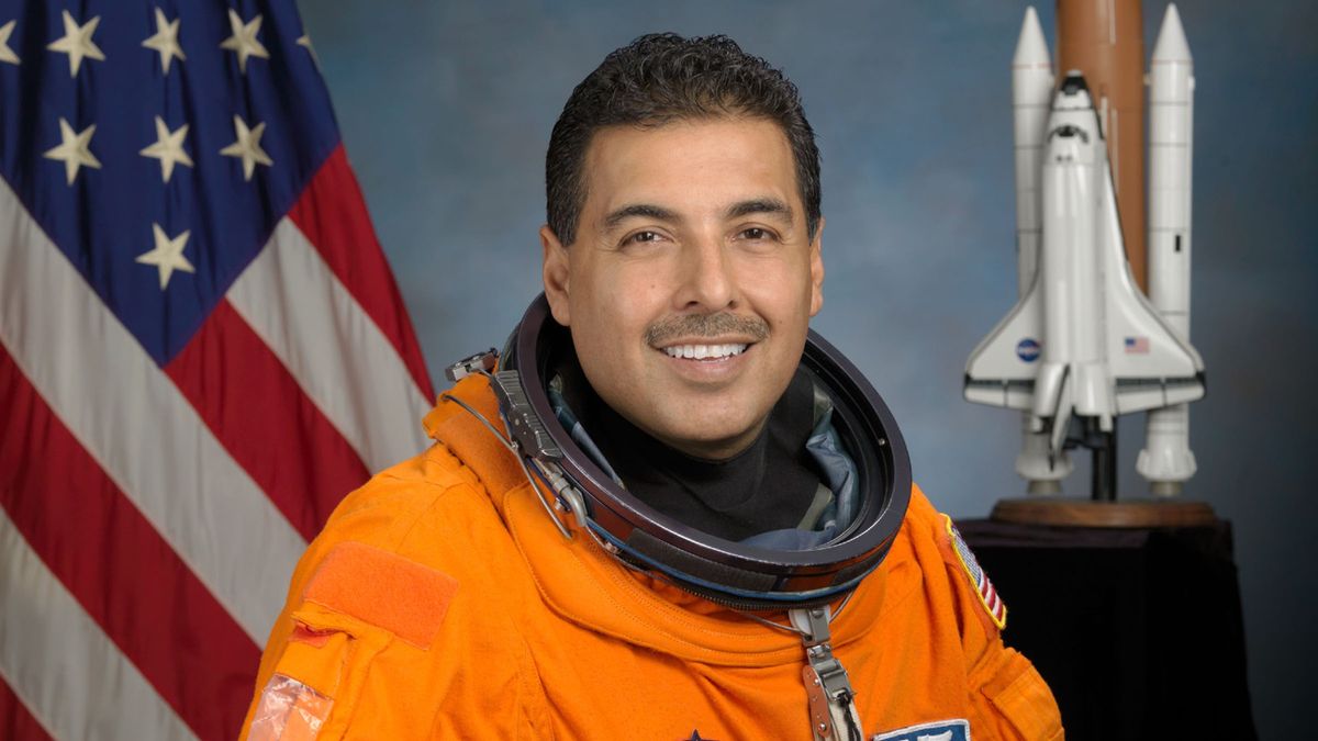 an astronaut in an orange spacesuit poses for a portrait in front of a model of a space shuttle