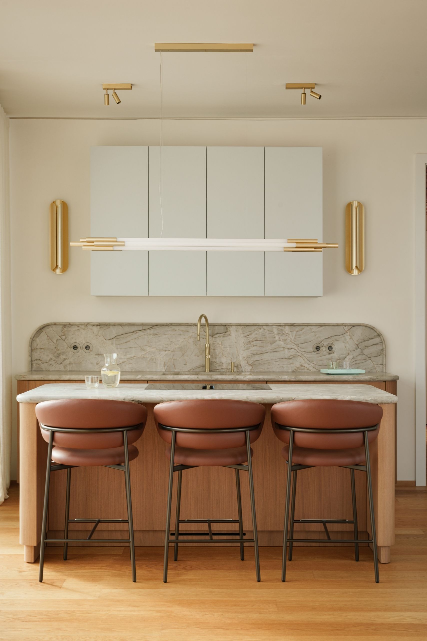 Image of a cream kitchen with a wooden island that has a green-tinted marble top, and three brown leather bar stools. There is light blue cabinetry above the sink and gold hardware.