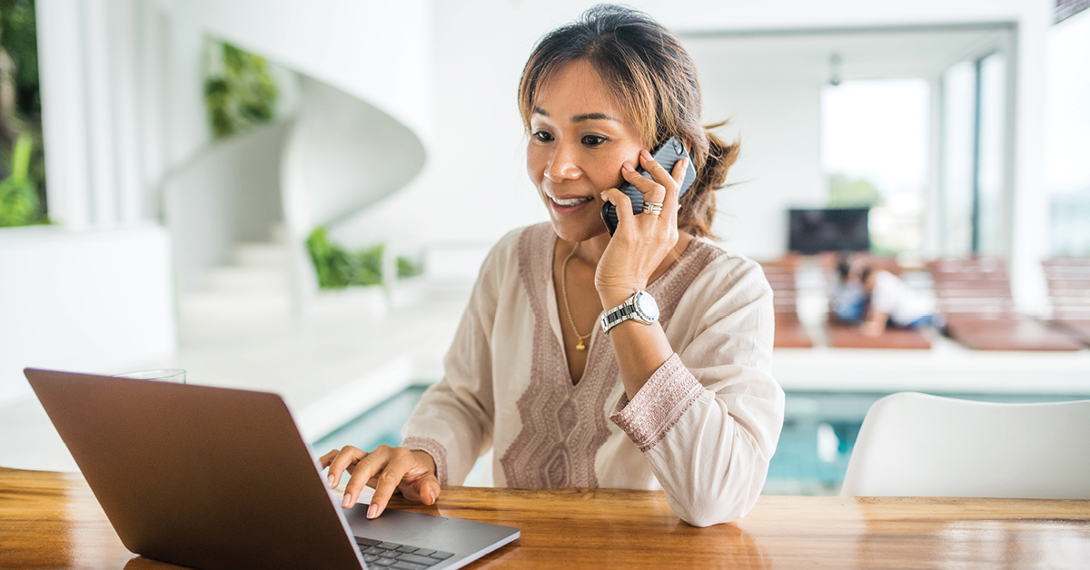 Asian woman on the phone sitting at a table with her laptop