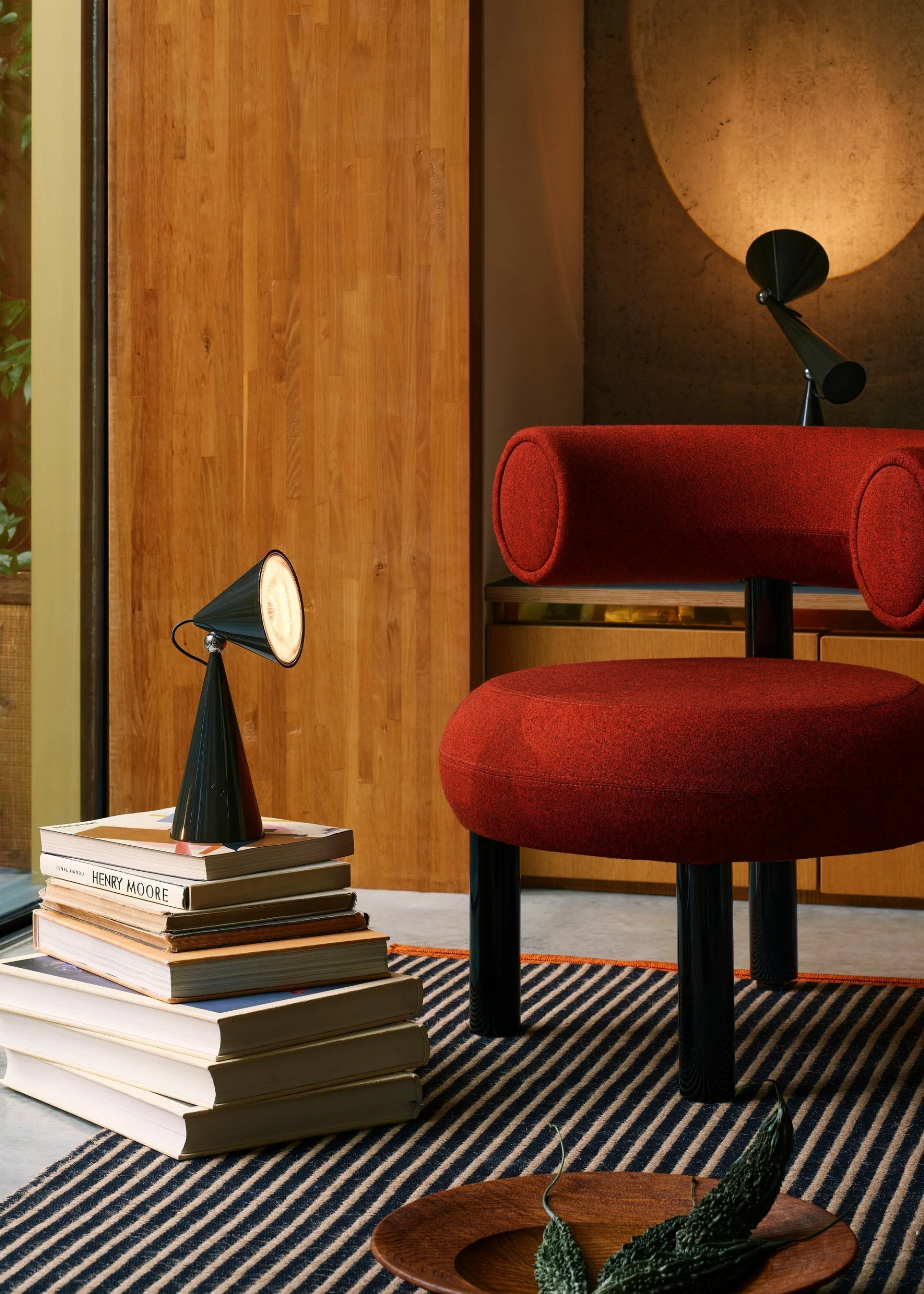 A living room with a geometric red chair, a stack of books and a table lamp on a striped rug, near a plate of vegetables