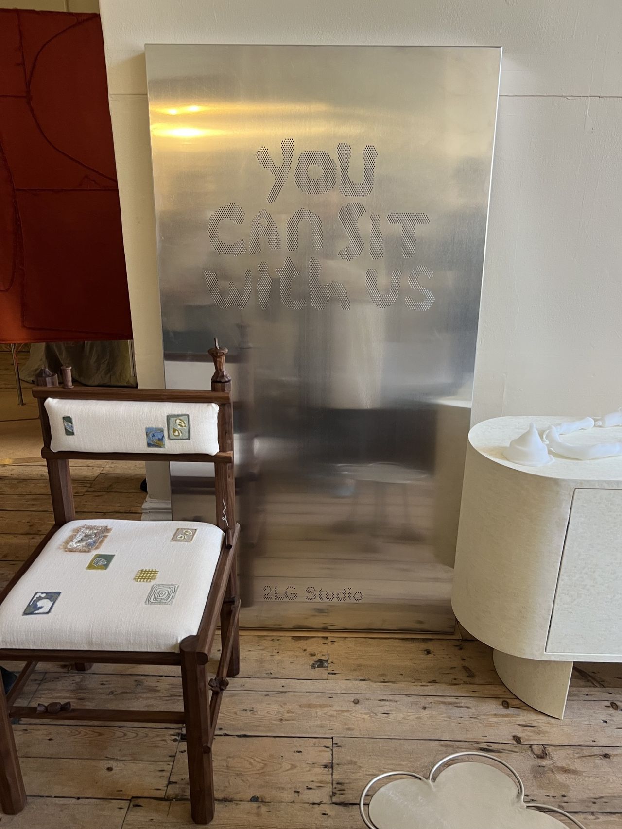 Image of a gallery with a silver sign that reads "you can sit with us", a wooden chair with stitching on the seat, and wooden floors.