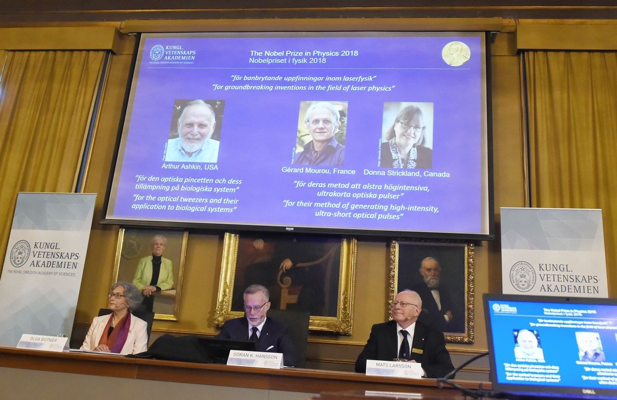 Members of the Nobel Committee for Physics sit in front of a screen displaying portraits of this year&#039;s Laureates: Arthur Ashkin, Gerard Mourou and Donna Strickland.