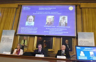 Members of the Nobel Committee for Physics sit in front of a screen displaying portraits of this year's Laureates: Arthur Ashkin, Gerard Mourou and Donna Strickland.