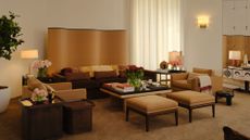 Neutral, richly layered living room featuring a wavy room divider, pair of ottomans, leather-topped coffee table, low-slung chocolate brown couch, softly aglow with ambient lighting