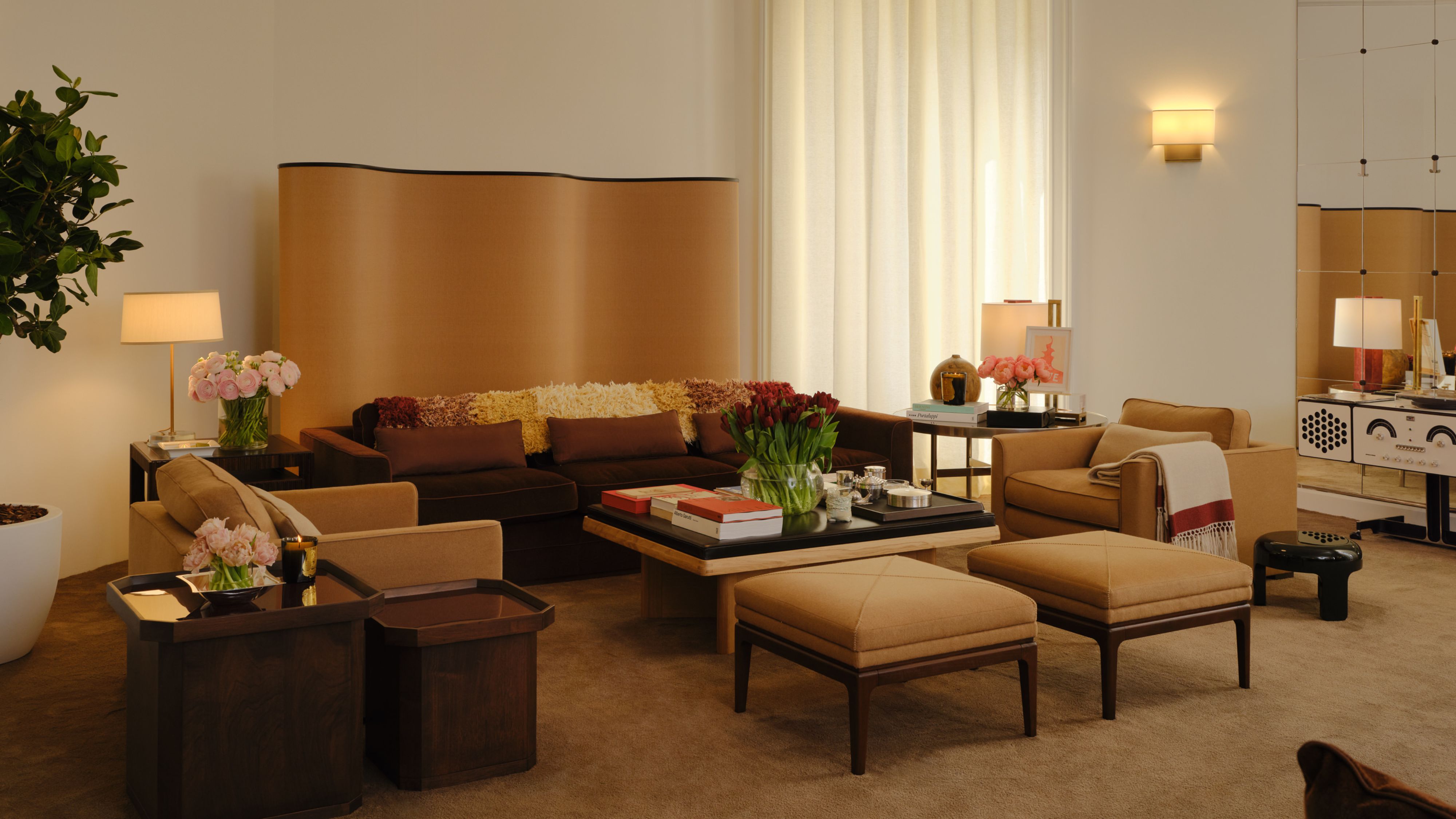 Neutral, richly layered living room featuring a wavy room divider, pair of ottomans, leather-topped coffee table, low-slung chocolate brown couch, softly aglow with ambient lighting