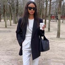 fashion influencer Lorna Humphrey of Symphony of Silk poses in a wooded park wearing black oversize sunglasses, a black blazer, white t-shirt, white trousers, and a black Prada bag