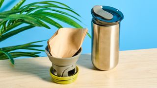 Wacaco Cuppamoka, a stainless steel coffee pour-over brewer, photographed against a blue background, with brown filters and a sippy lid