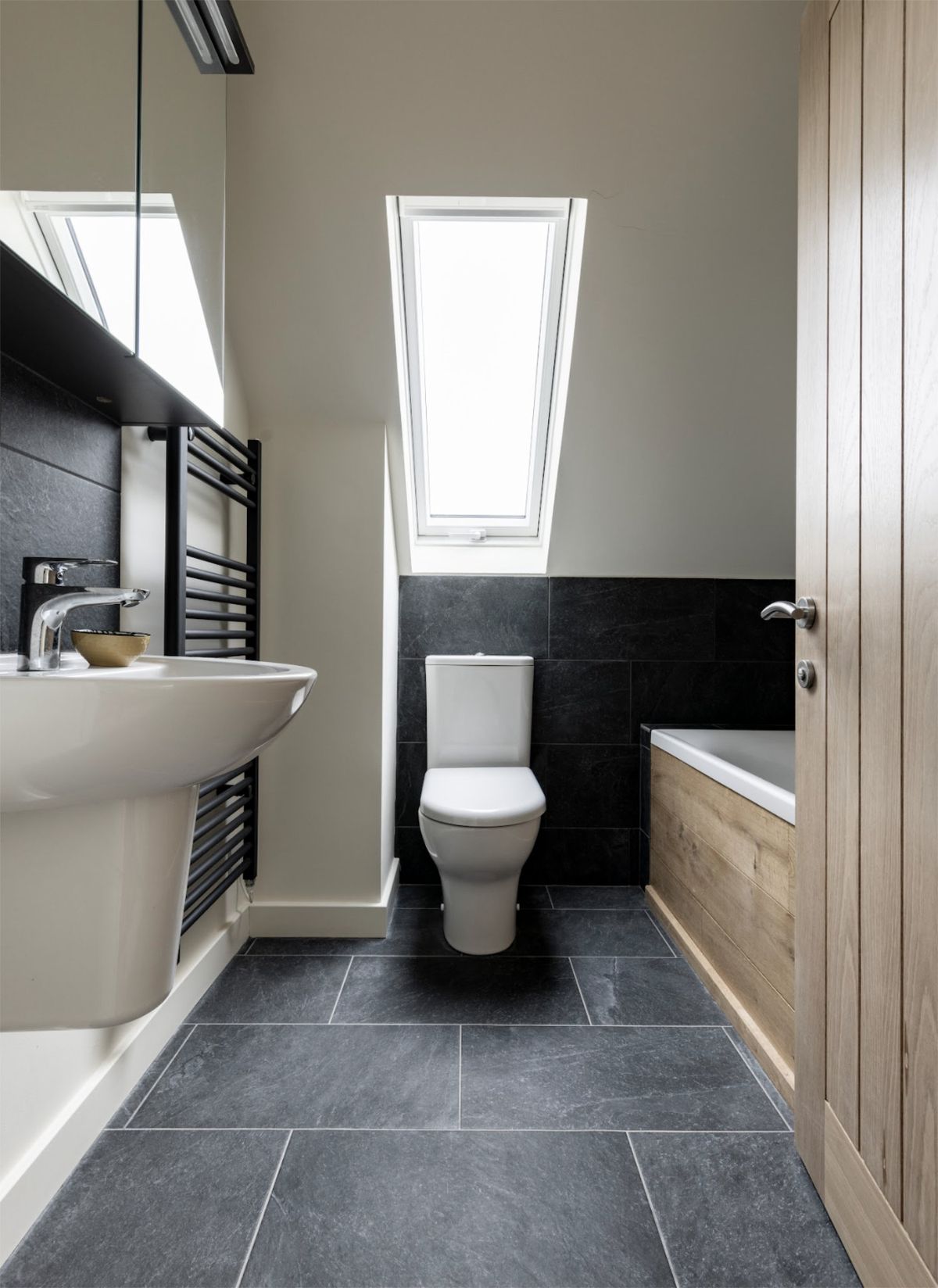 A small bathroom with a dark tiled floor, a heated towel rail and a wooden panelled bathtub