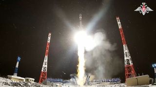 A white and green rocket launches at night surrounded by falling snow and red and white towers