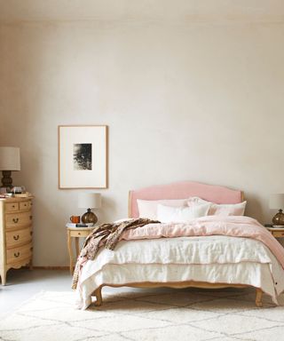A bedroom with beige walls, a light pink bed frame with white ruffled bedding, a wooden set of drawers to the left and a white and black art print on the wall