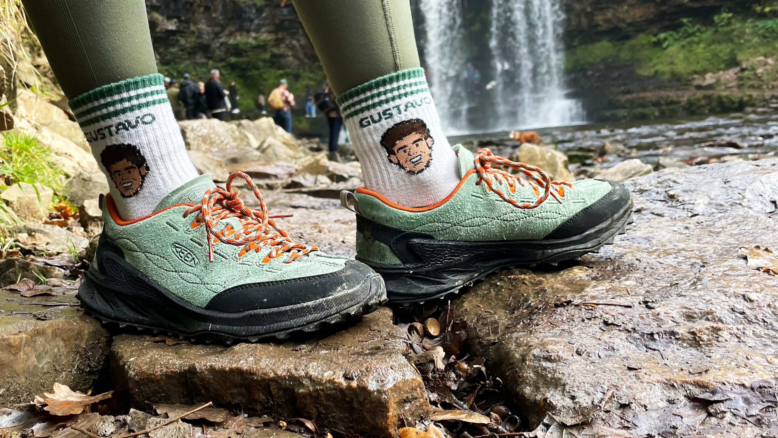 A picture of the Keen Jasper Zionic hiking shoes in green/gold colorway being worn on a hiking trail with a waterfall in the background