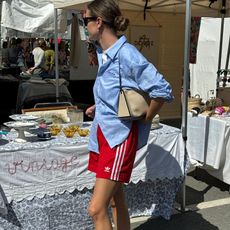 French woman wears red Adidas three stripe shorts, a blue button down and Chanel flats at an outdoor market.