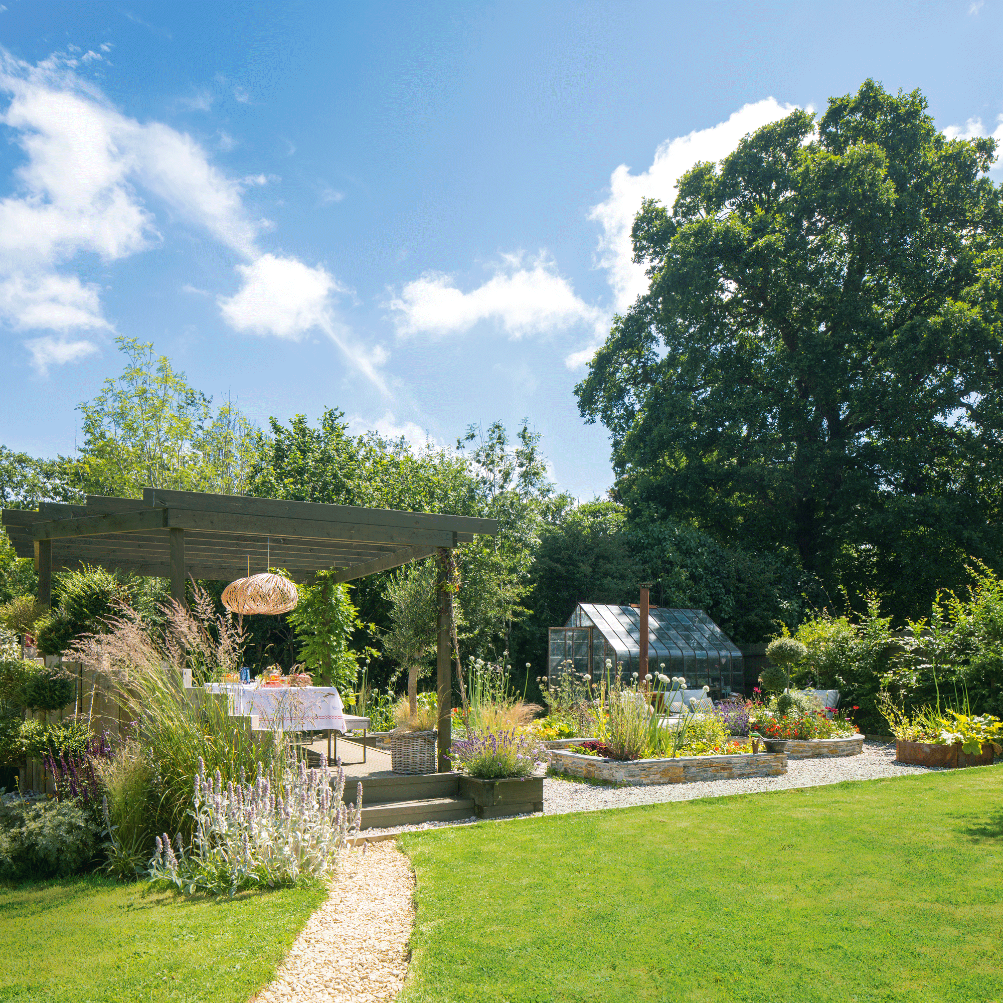 a garden with a lawn with a pebbled pathway through the middle leading to a pergola covered dining area and greenhouse
