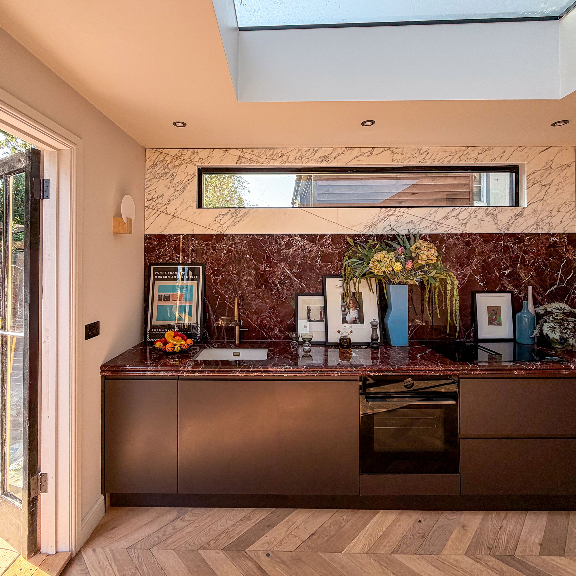 Kitchen with wooden chevron flooring, red marble worktops and splashbacks, topped with white marble. Dark red cabinetry. High narrow window. Photographs and vases on countertop. Skylight in ceiling.