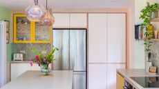 Kitchen color ideas are so chic. Here is a kitchen with a white island and cabinets, green and white walls, a silver fridge, yellow lower cabinets, and iridescent pendant lights