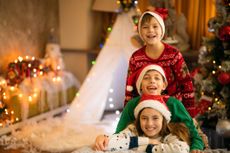 Three children, probably siblings, lined up wearing Christmas sweaters and hats in front of a Christmas tree.