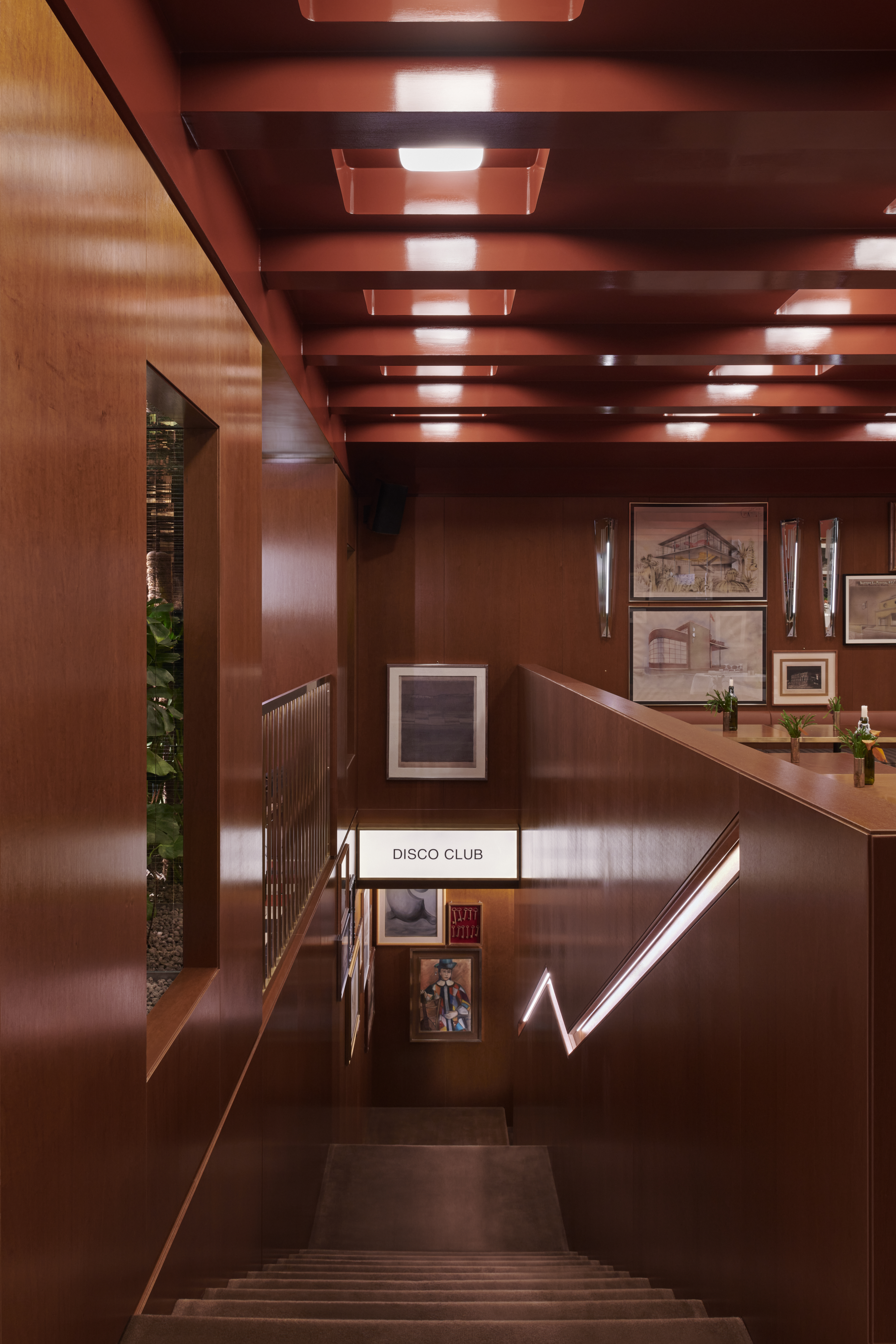 A restaurant&#039;s dining room boasts back-lit coffered ceilings, mid-century furniture in leather, chrome, and wood, framed artworks scattered across its walls, checkered floors, and a downstairs bar clad in brushed steel and golden light.