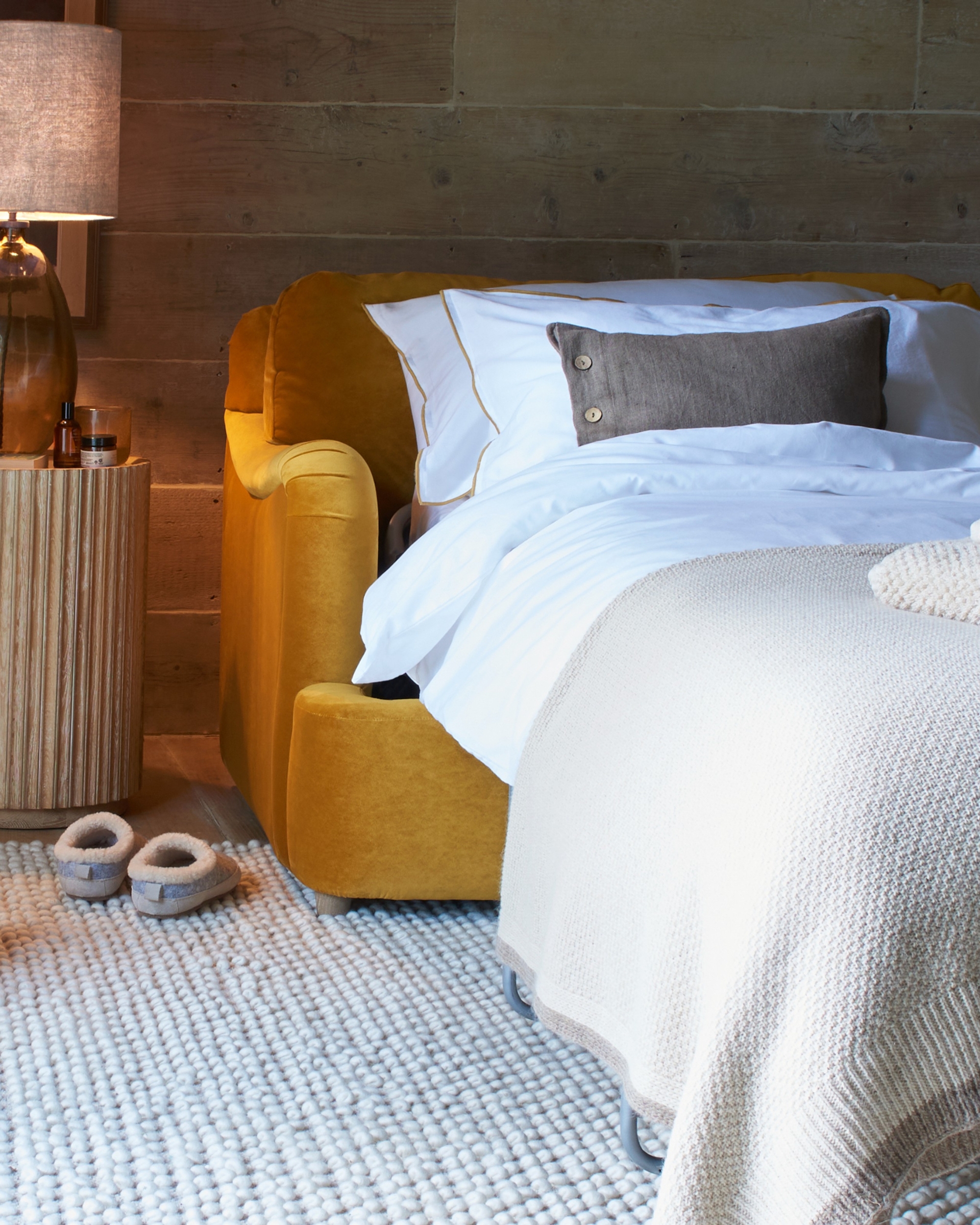 Image of an ochre yellow love seat sofa bed with the mattress pulled out and made up. There is a knotted rug on the ground and slippers by the bed.